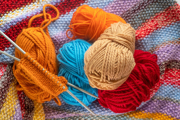 Pelotes de laine colorées (orange, bleue, beige, rouge) et aiguilles à tricoter posées sur un tissu tricoté multicolore - Agrandir l'image, fenêtre modale