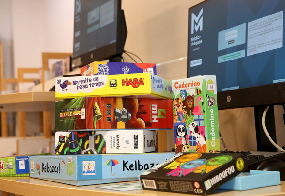 Boîtes de jeux empilées sur un comptoir de médiathèque : Kelbazar, Cadomino, Meeple Hope, Tantrix et Hiboufou. - Agrandir l'image, fenêtre modale