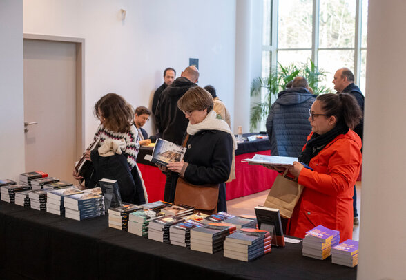 Des visiteurs consultent et choisissent des ouvrages parmi de nombreuses piles de livres disposées sur des tables noires. - Agrandir l'image, fenêtre modale