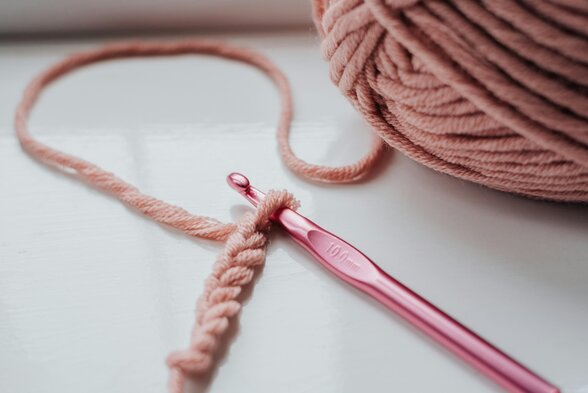 Un crochet rose métallique engagé dans une chaînette de mailles, posé près d'une pelote de fil vieux-rose - Agrandir l'image, fenêtre modale