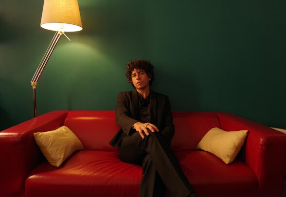 Brian López, songwriter et guitariste, assis sur un canapé rouge dans une ambiance feutrée et colorée - Agrandir l'image, fenêtre modale
