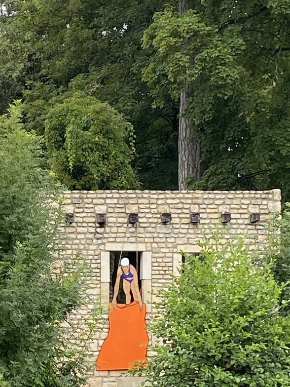Installation Les Baigneuses d'Anne Vergneault : performeuse en maillot brandissant un tissu orange depuis une bâtisse en pierre - Agrandir l'image, fenêtre modale