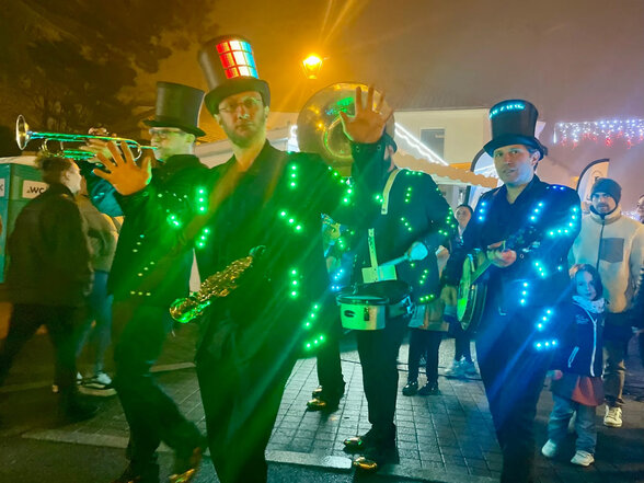 Musiciens en costume noir orné de LED colorées et hauts-de-forme, jouant trompette et saxophone de nuit en plein air devant des passants - Agrandir l'image, fenêtre modale