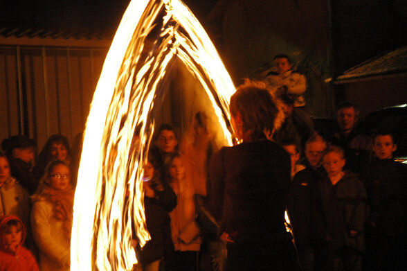 Une artiste fait tournoyer un large arc de flammes devant un cercle de spectateurs, adultes et enfants, lors d'une soirée en plein air. - Agrandir l'image, fenêtre modale