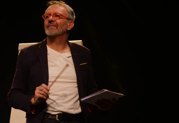 Jérôme Rouger en scène, baguette et carnet en main, regard levé vers le public avec un sourire complice - Agrandir l'image, fenêtre modale