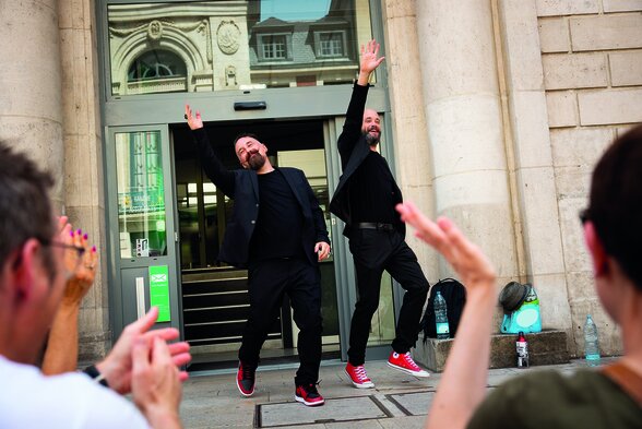 Deux comédiens en noir aux baskets rouges saluent bras levés un public qui applaudit, lors d'une représentation en plein air - Agrandir l'image, fenêtre modale