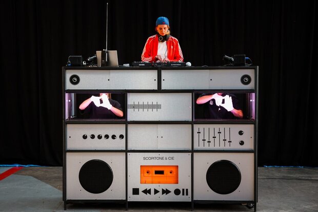 Une DJ mixe depuis un pupitre scénographique géant en forme de ghetto blaster orné d'une cassette orange et d'écrans intégrés