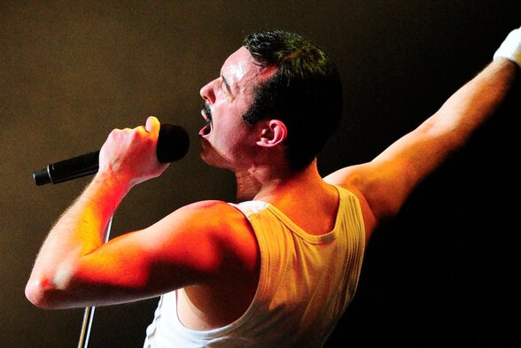Gary Mullen incarne Freddie Mercury sur scène, micro en main et bras levé, sous les feux des projecteurs - Agrandir l'image, fenêtre modale