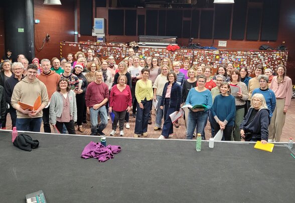 Une cinquantaine de choristes posent ensemble sur scène, partitions en main, dans une salle de concert équipée de projecteurs. - Agrandir l'image, fenêtre modale