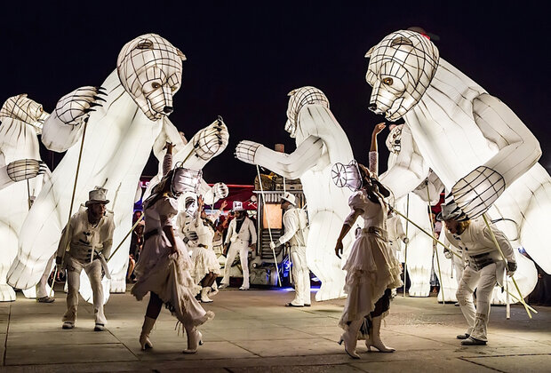 Marionnettes géantes d'ours lumineux blancs encadrant des performeurs en tenues blanches lors d'un spectacle nocturne en plein air
