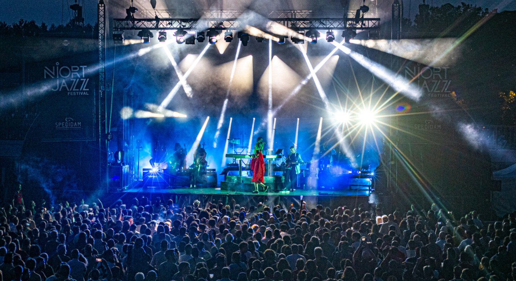 Grande scène illuminée de faisceaux bleus, artiste en tenue rouge face à une foule nombreuse lors d'un concert en plein air
