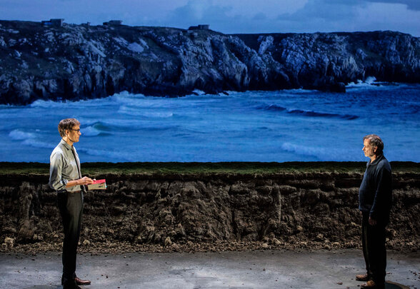 Deux comédiens face à face sur un plateau nu, devant une projection de côte rocheuse battue par les vagues. - Agrandir l'image, fenêtre modale