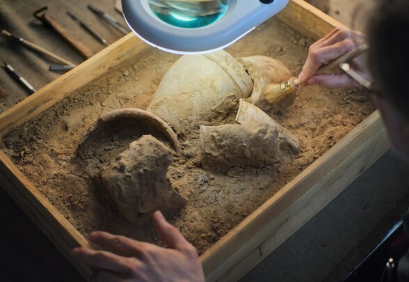 Restaurateur nettoyant au pinceau des fragments de poterie ancienne dans un bac de sable, sous une loupe éclairante - Agrandir l'image, fenêtre modale