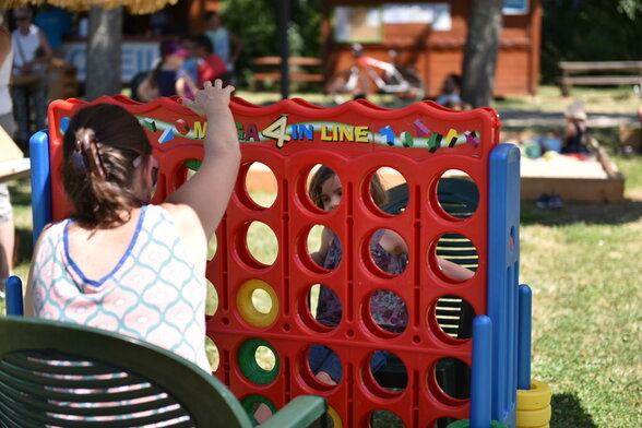 Participante glissant un palet dans un Méga 4-en-ligne géant en plein air, un enfant visible à travers la grille colorée. - Agrandir l'image, fenêtre modale