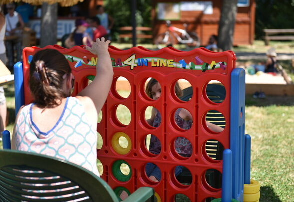 Participante glissant un palet dans un Méga 4-en-ligne géant en plein air, un enfant visible à travers la grille colorée. - Agrandir l'image, fenêtre modale