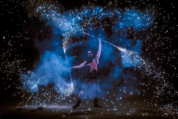 Artiste brandissant des bâtons pyrotechniques au cœur d'une nuée d'étincelles et de fumée bleue nocturne