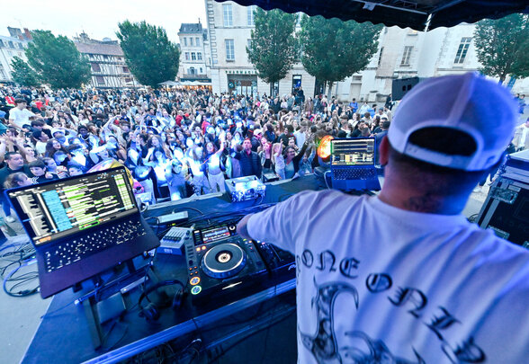DJ mixant depuis une scène en plein air, de dos face à une foule dense rassemblée sur une place du centre-ville - Agrandir l'image, fenêtre modale