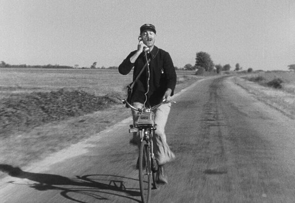 Photogramme en noir et blanc : le facteur François à vélo sur une route de campagne, téléphone à l'oreille - Agrandir l'image, fenêtre modale