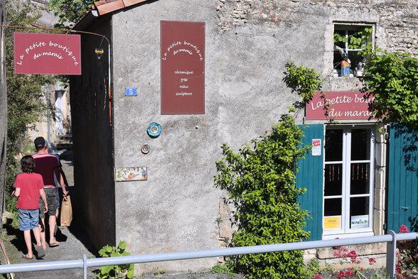 Façade de pierre avec l'enseigne 'La petite boutique du marais' et deux visiteurs s'engageant dans la ruelle - Agrandir l'image, fenêtre modale