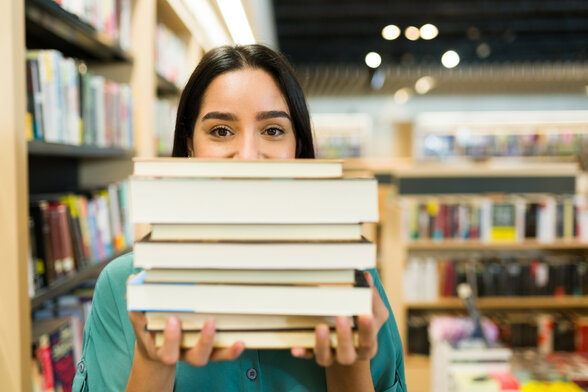 Une femme tient une pile de livres devant son visage en souriant, dans une librairie aux rayonnages bien garnis - Agrandir l'image, fenêtre modale
