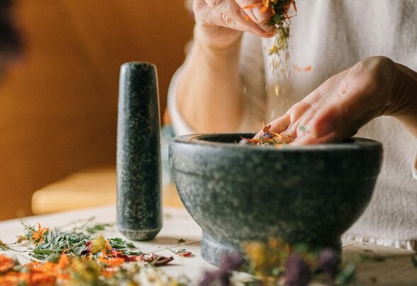 Mains versant des fleurs et herbes séchées dans un mortier en pierre, sur une table parsemée de plantes colorées - Agrandir l'image, fenêtre modale