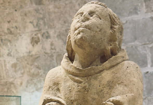 Statue médiévale en pierre d'un personnage religieux en robe, les bras levés, exposée dans une salle aux murs de pierre - Agrandir l'image, fenêtre modale