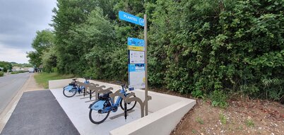 Station VLS équipée de deux vélos garés, borne d'information et panneau indicateur « Plantes », en bordure de route