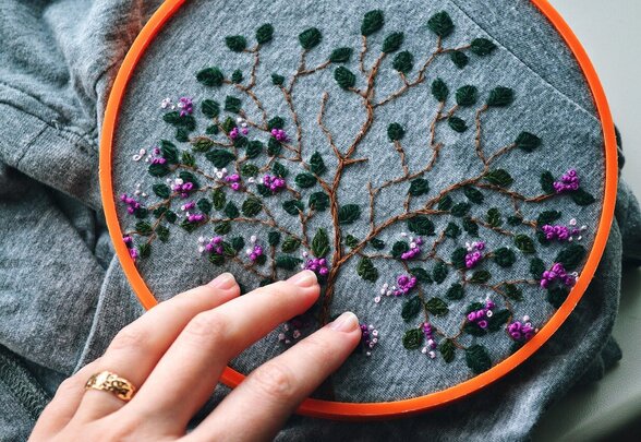 Main posée sur un tambour à broder orné d'un arbre aux feuilles vertes et petites fleurs violettes - Agrandir l'image, fenêtre modale