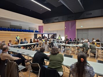 Réunion en salle avec plusieurs dizaines de participants assis aux tables, face aux intervenants au fond